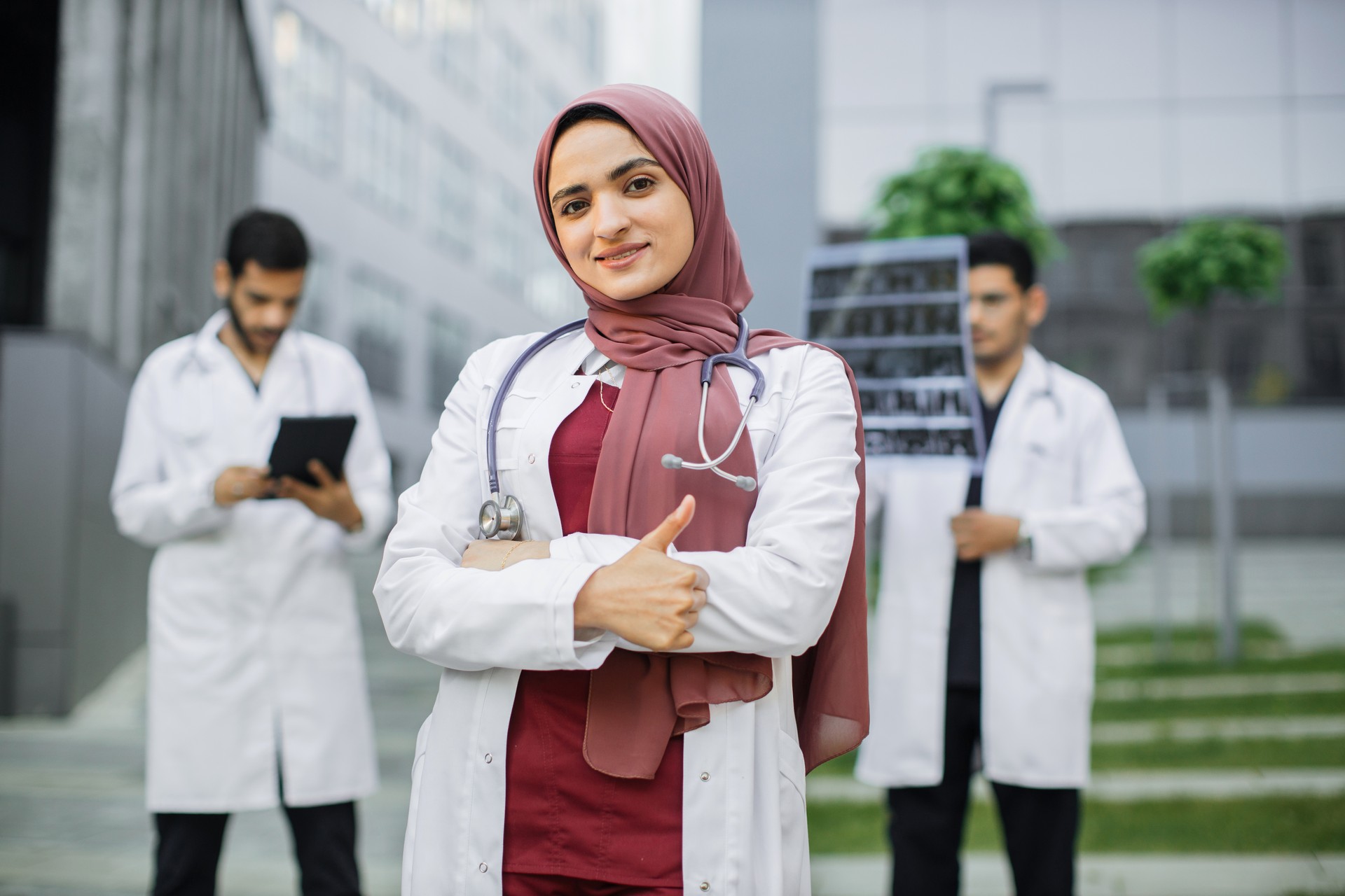 Female smiling Arab doctor in hijab showing thumb up while her two male colleagues standing behind Female smiling Arab doctor in hijab showing thumb up while her two male colleagues standing behind